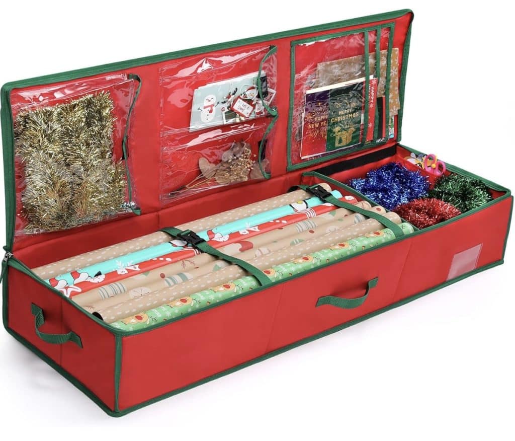 A red storage box is open, revealing compartments filled with colorful wrapping paper rolls, tinsel, and ribbons alongside cute Christmas tags. Transparent pockets on the lid hold various small wrapping items. The box is accented with green handles and trim.